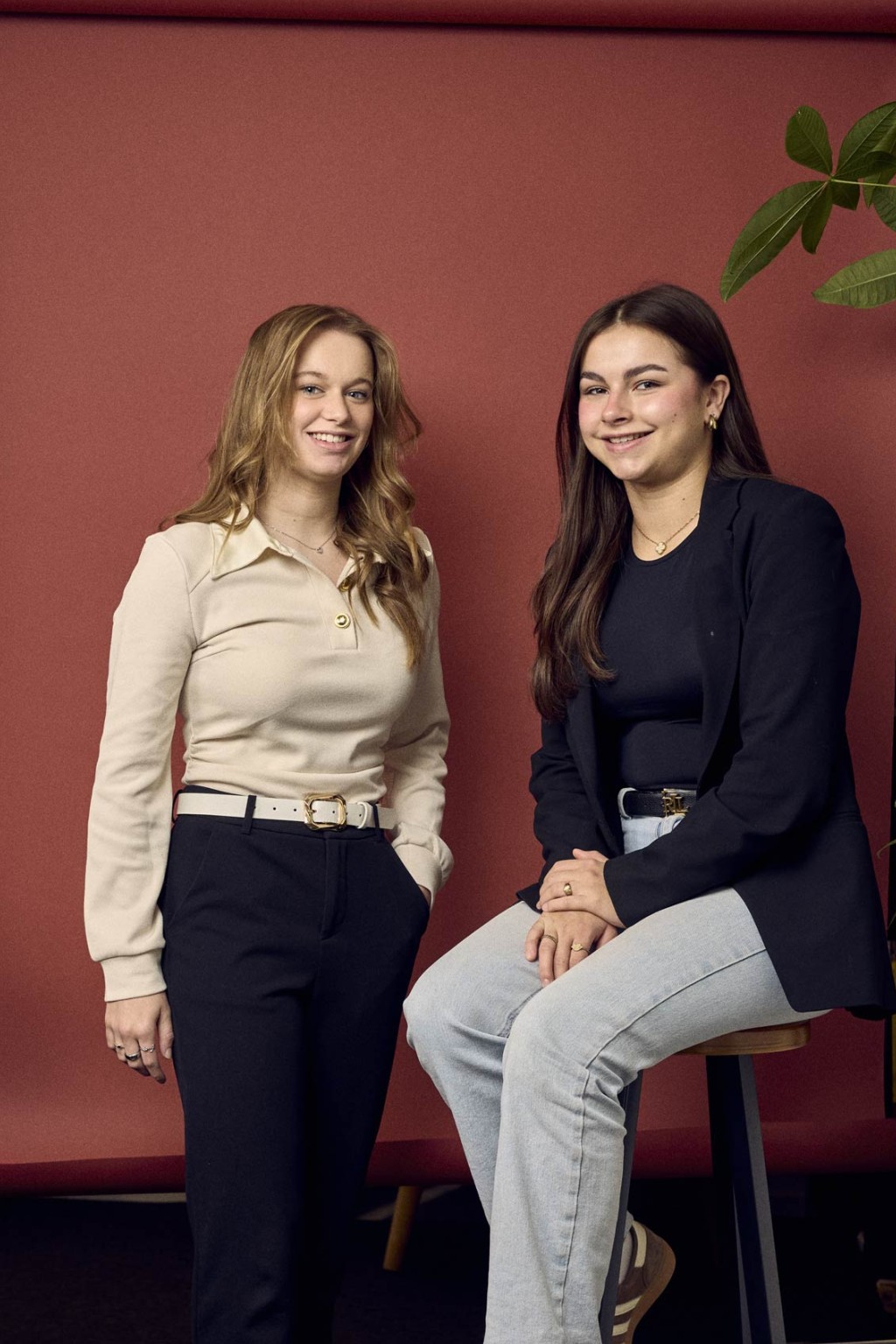 Amy Cluitmans en Isis van Heugten poseren samen voor een rode foto backdrop in kantoor, een iemand zit op een kruk en een iemand staat er langs.