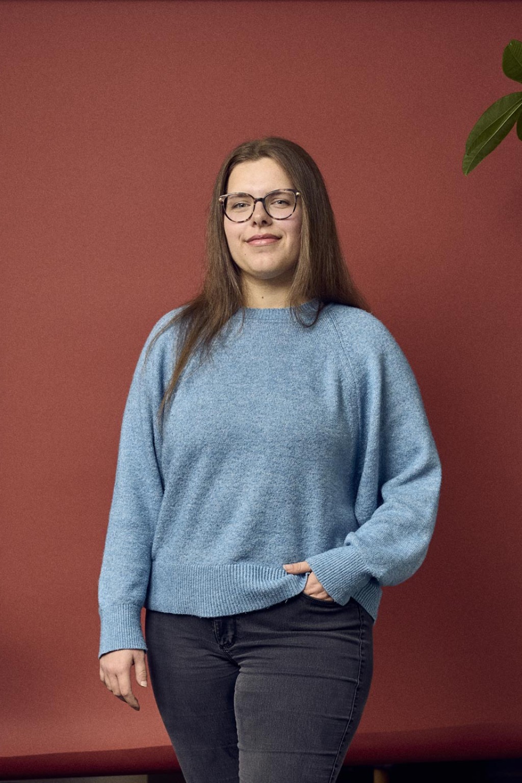 Chayenne Noben staat in een lichtblauwe trui voor een rode backdrop met een plant langs haar in een kantoorruimte.