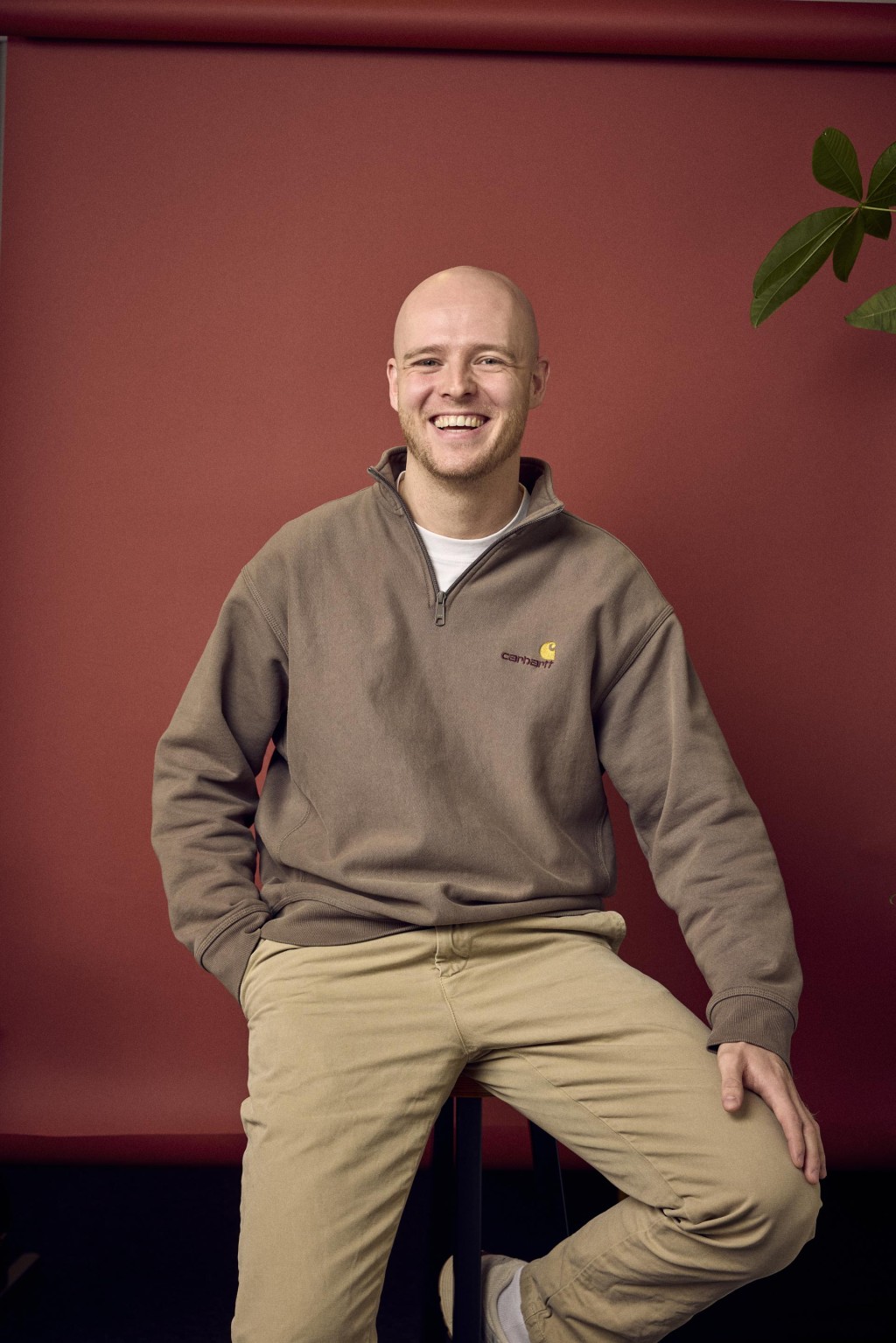 Jeffrey Caris zittend op een kruk voor een rode backdrop met een bruine trui aan.