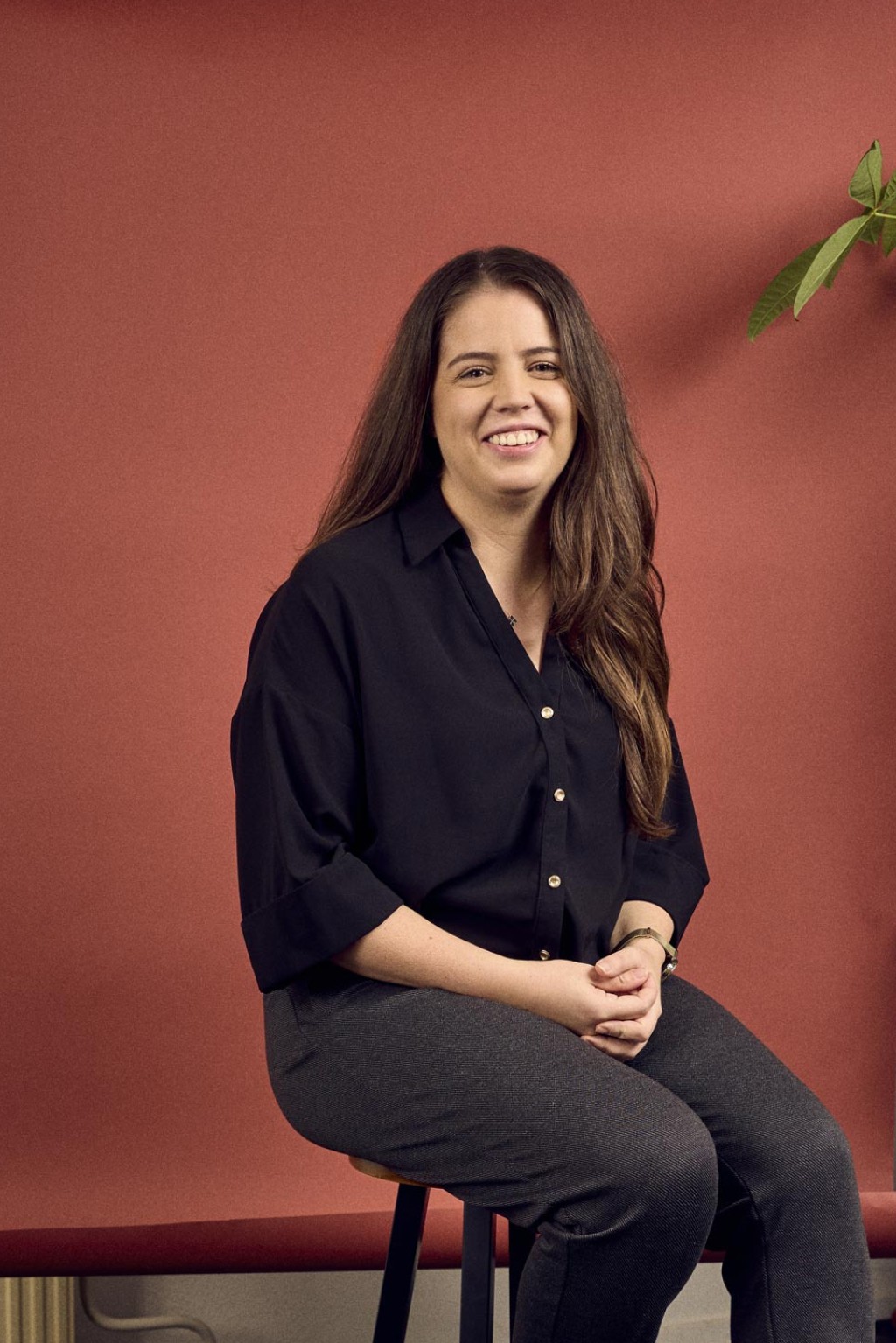 Kelly Reijnders zittend op een kruk voor een rode backdrop in een kantoorruimte met een plant rechts van haar.