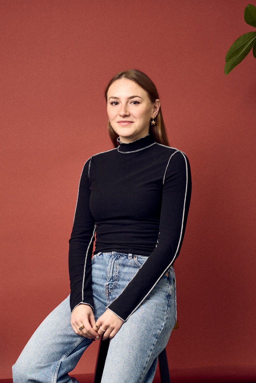 Lieke van Engelenhoven zit op een kruk voor een rode foto backdrop in een kantoorruimte met een zwarte lange mouw top aan.