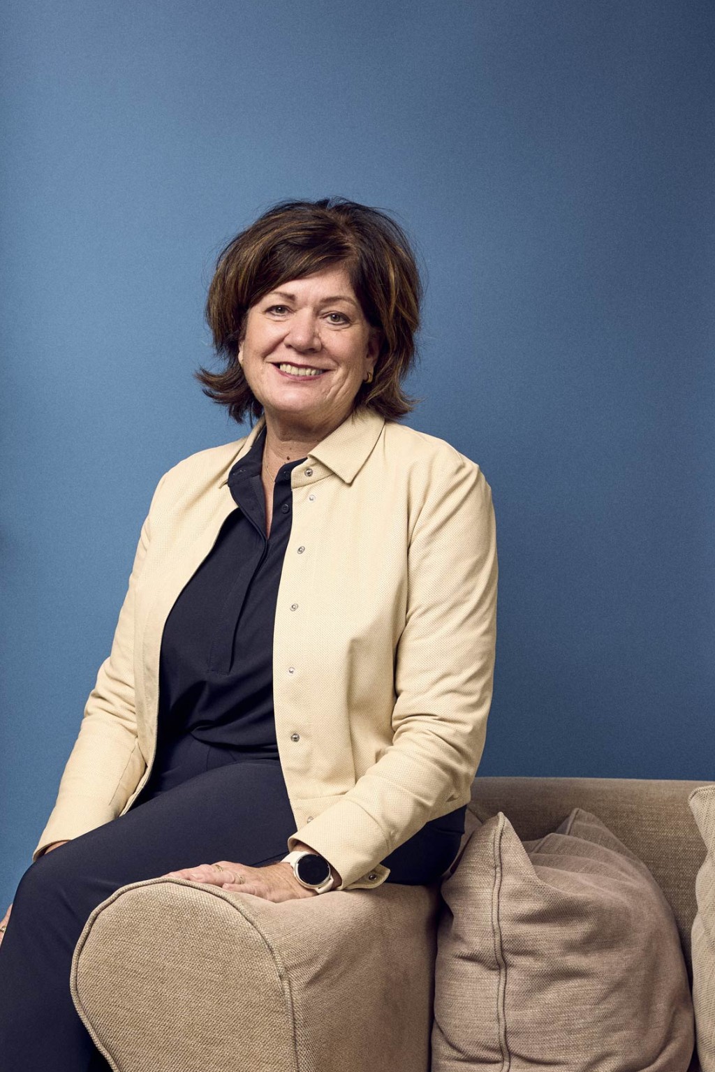 Monique Dekkers in een kantoorruimte met een blauwe backdrop zittend op een leuning van de bank.