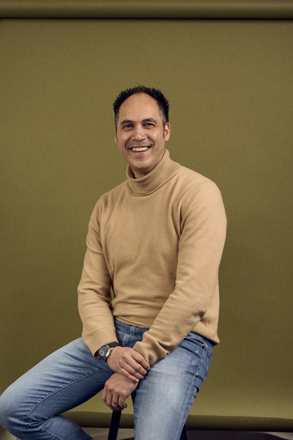 Owen Manusama zittend op een kruk voor een groene backdrop in een kantoorruimte.