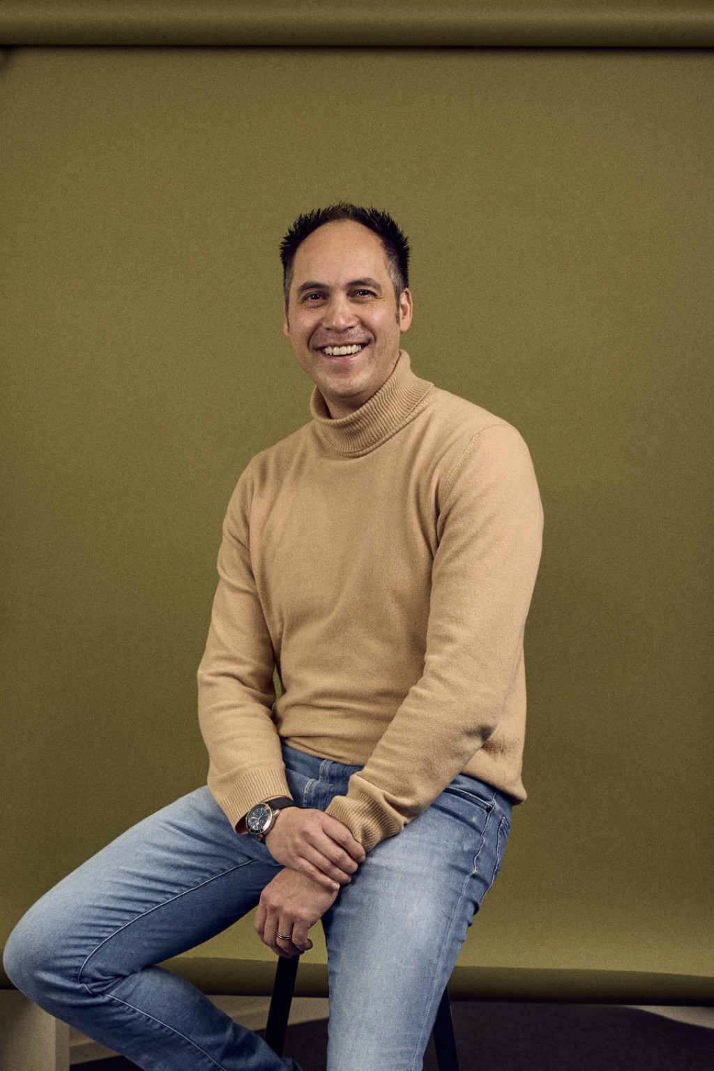 Owen Manusama zittend op een kruk voor een groene backdrop in een kantoorruimte.