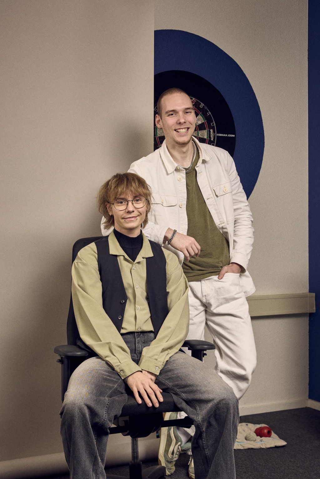 Rivian Vissers en Xander Kuijpers staan naast elkaar voor een bronzen backdrop, één zittend op een kruk en één staand, beiden lachend naar de camera.