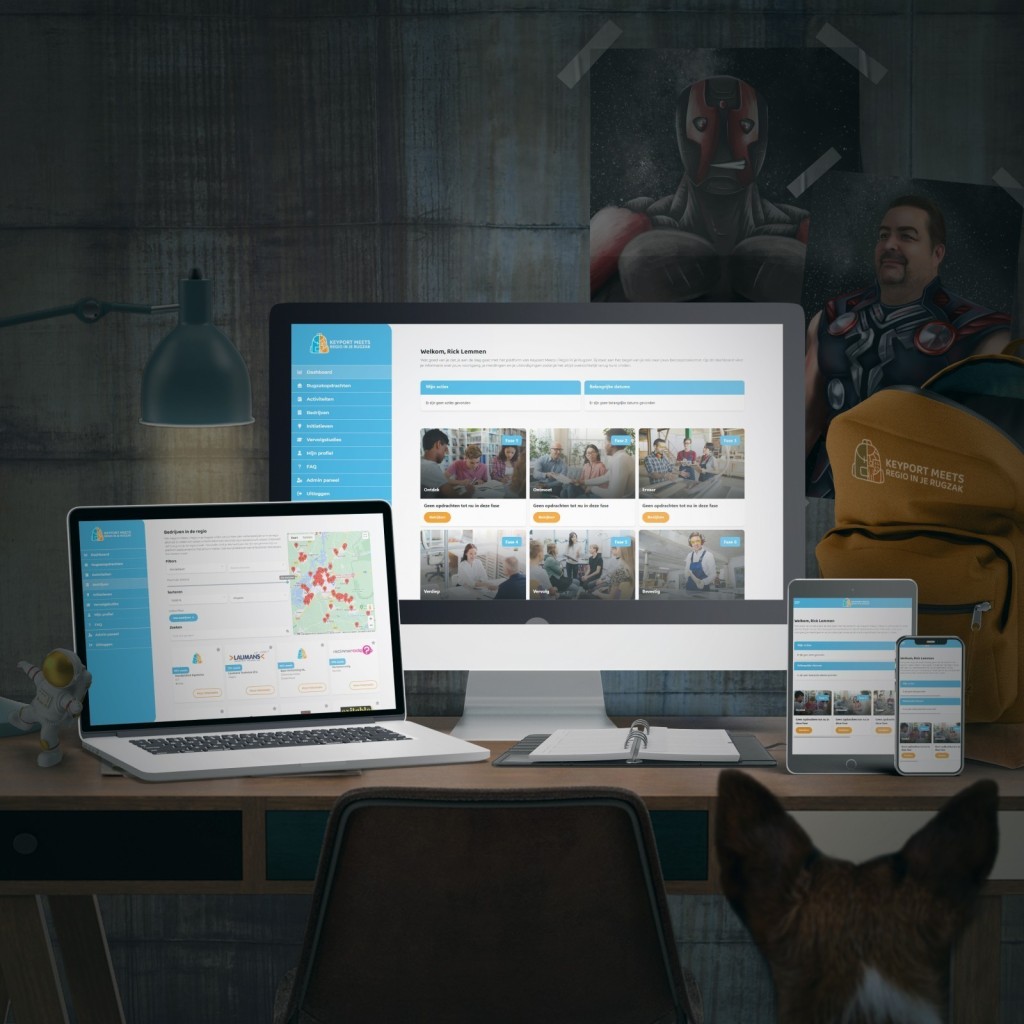 Bureau met daarop verschillende apparaten waarop de website van Regio in je Rugzak staat, met op de achtergrond de Rugzak en twee superhelden posters.