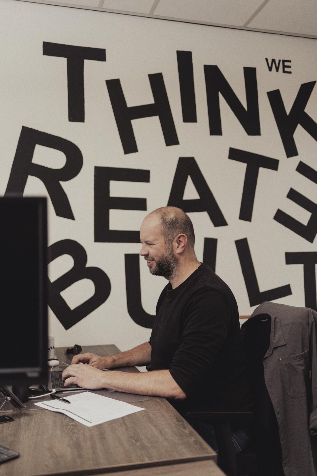 Een collega zit te werken aan een bureau, met op de achtergrond een grote muurtekst “Think, Create, Build”.