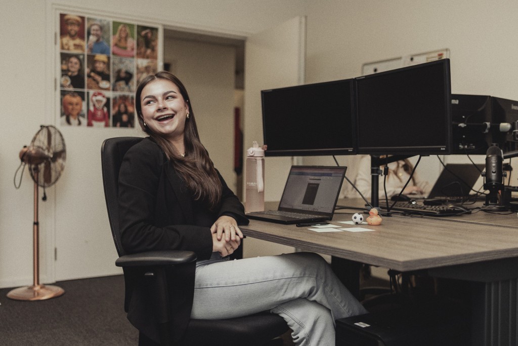 Een vrouw zit achter haar bureau met meerdere computer schermen en lacht naar haar collega achter haar.