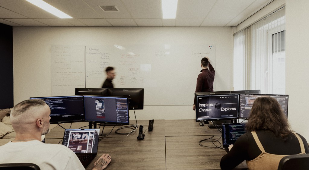 Twee mensen kijken naar twee grote computerschermen met code, terwijl een derde collega op een bord schrijft en iemand anders door de ruimte loopt