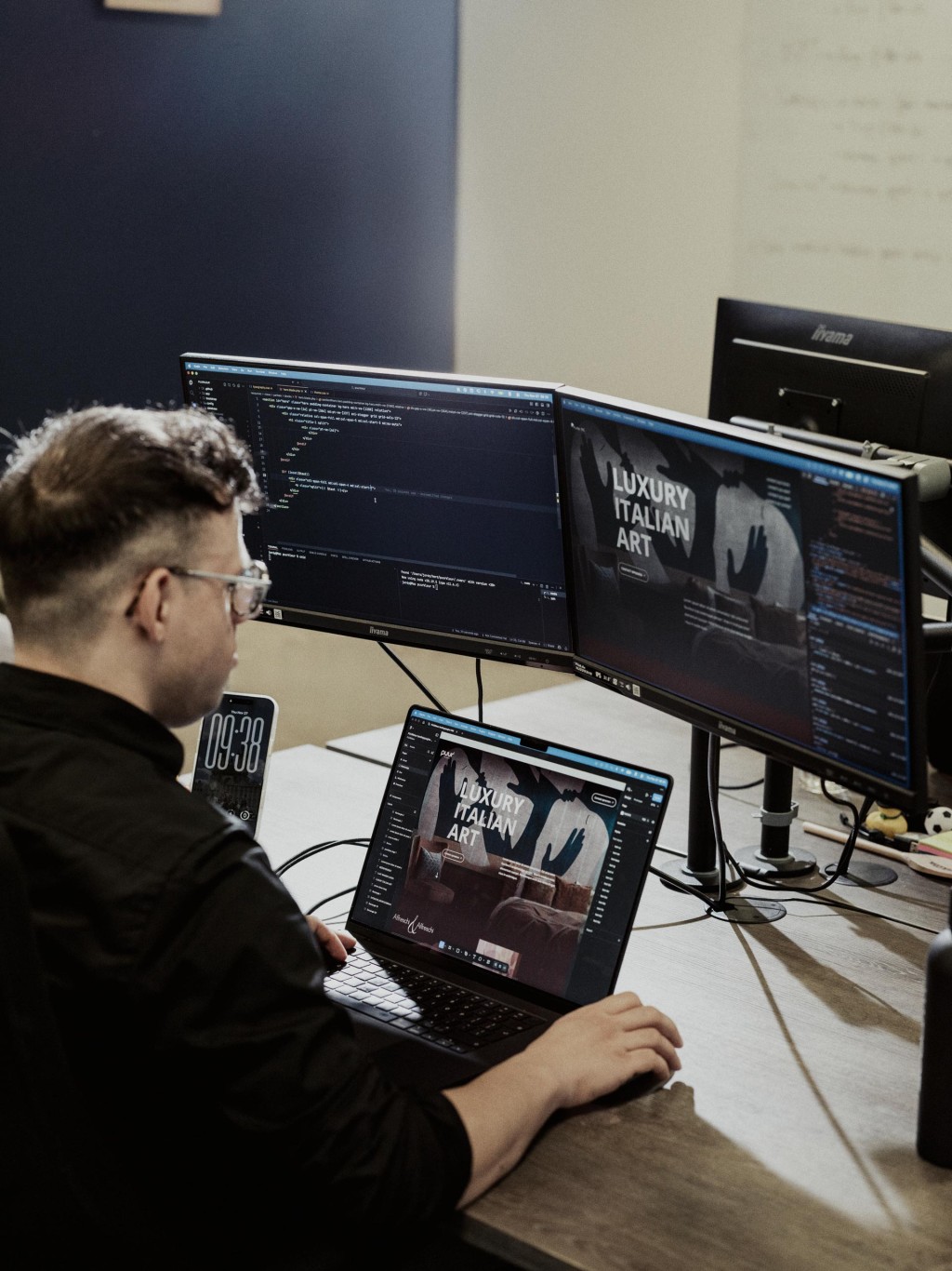 Een developer zit achter drie computerschermen waarop code en applicaties zichtbaar zijn