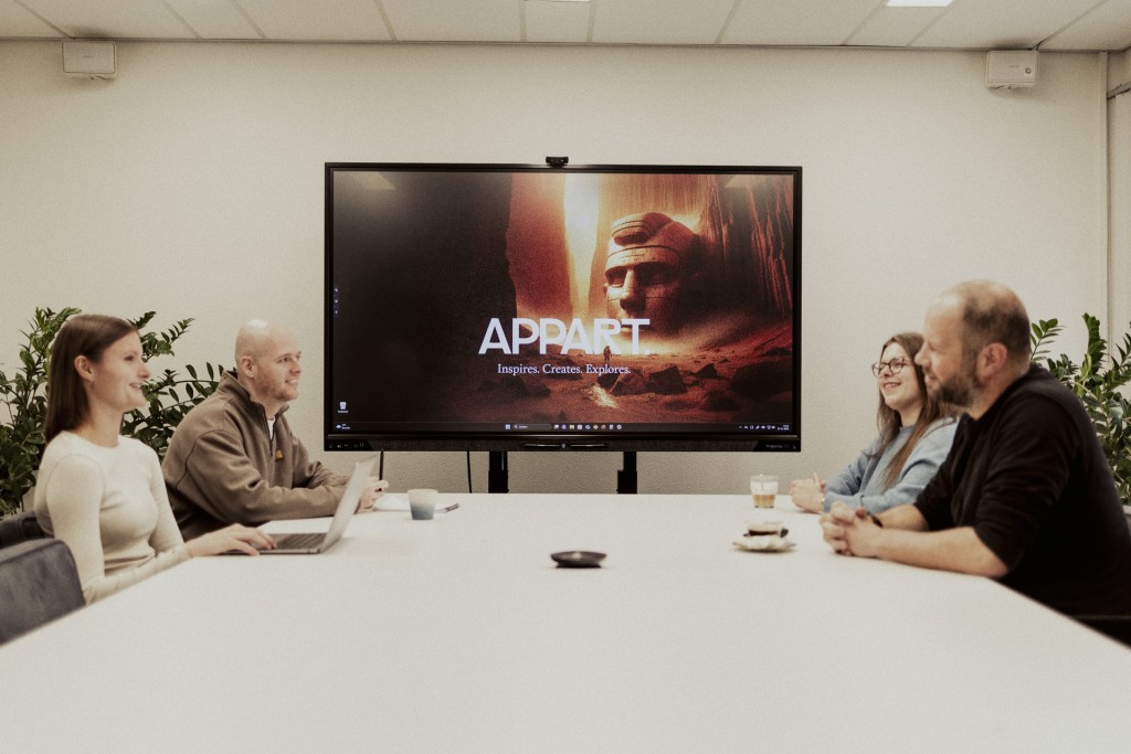Vier collega’s zitten aan een vergadertafel terwijl een groot scherm met het Appart logo achter hen hangt.