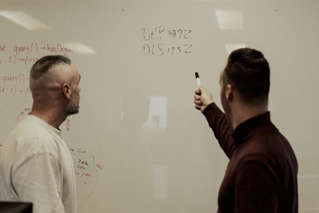 Twee mannen staan bij een whiteboard vol geschreven notities en bespreken de inhoud